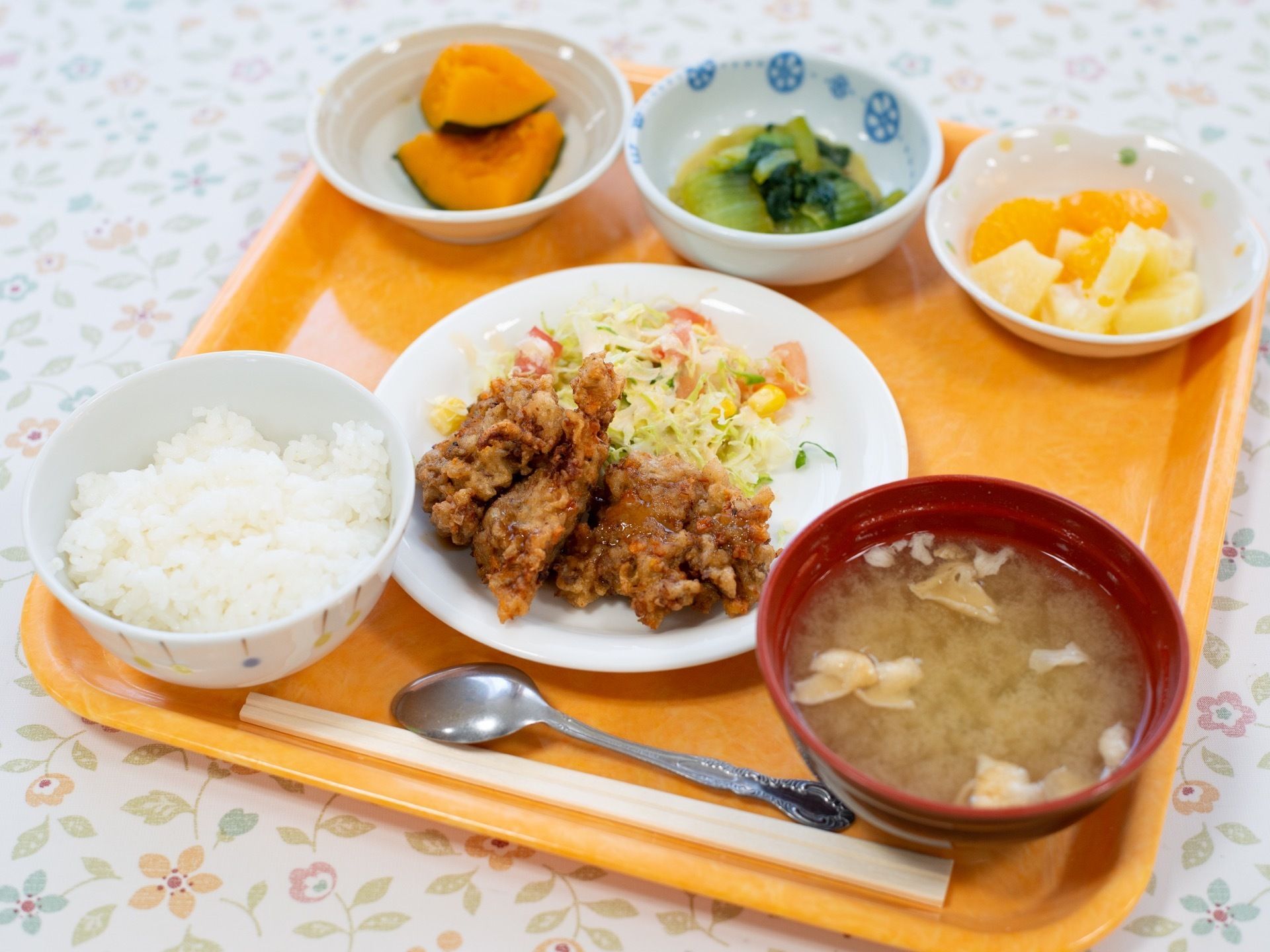四角いトレイの上に、ご飯と味噌汁、主菜、副菜２つと果物がある。栄養や彩りが偏らないようにバランスよく考慮されている。