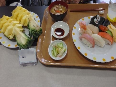 木製のトレイの上に握り寿司と醤油、白菜の漬物がそれぞれ大小の丸い皿に盛られ、汁物と箸置きに置かれた箸、コップも置かれている。トレイ左の皿には生のパイナップルが盛られている。