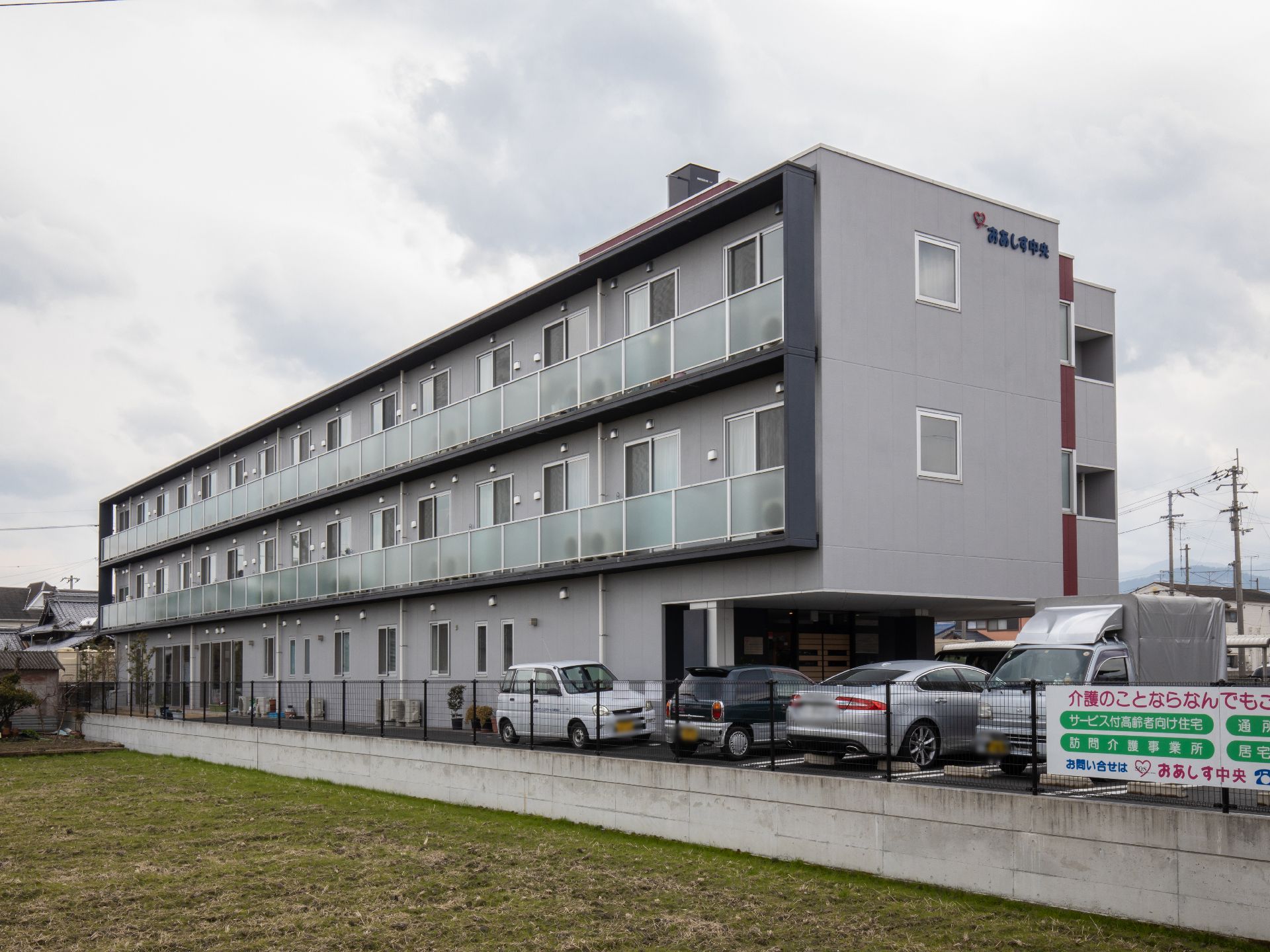 グレーの外壁の３階建ての建物で、壁面には窓が並んでいる。建物の横に駐車場があり、車が複数停まっている。