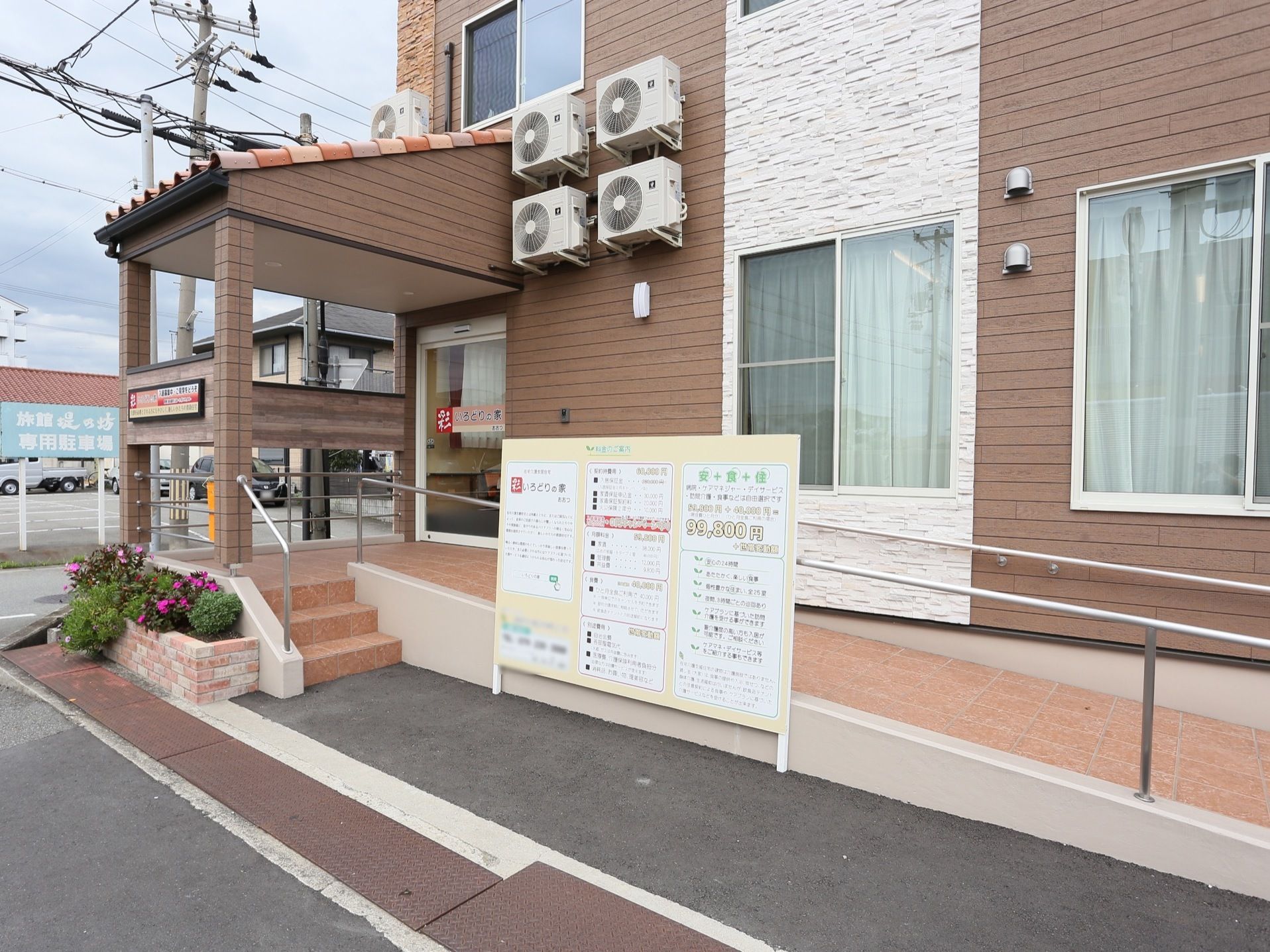 玄関までは長いスロープが設けられている。玄関前には花壇がある。施設や入居料金の案内がスロープの手前に設置されている。