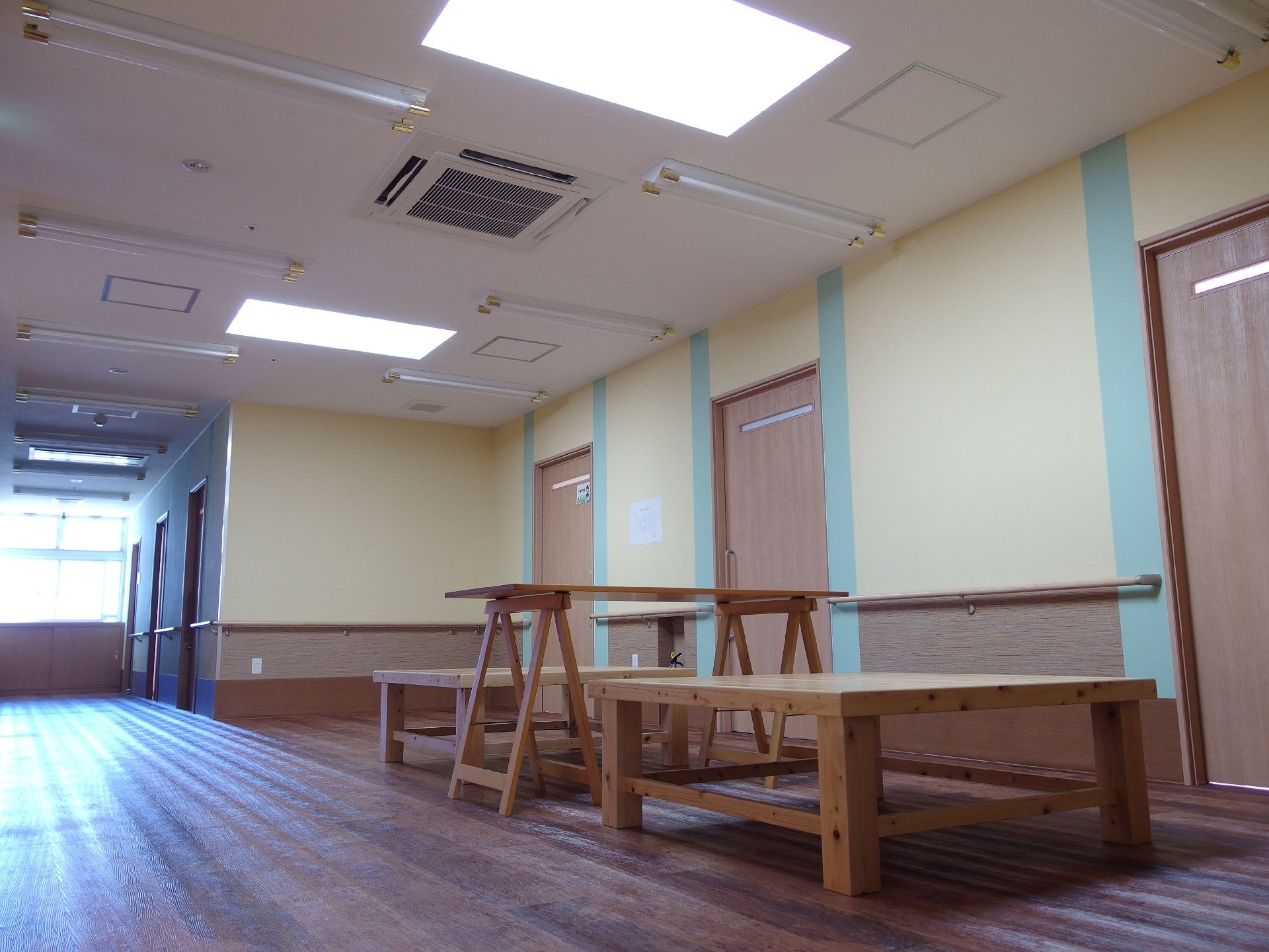 居室前に木製のベンチ二つと机。左手に廊下が伸びていて、突き当たりの窓から日差しが降り注いでいる。