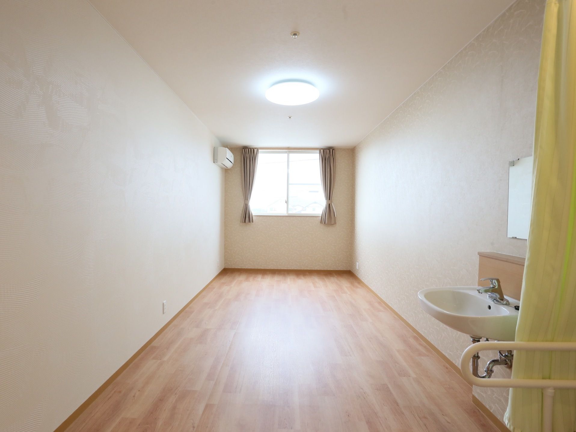 白い壁紙に木目が美しい床材が敷き詰められ、明るく開放的な部屋である。エアコンと洗面台が設置されている。
