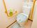 両サイドのオレンジの手すりと呼び出しコールが付いた居室トイレ。グリーンのトイレカバーが付けられ、トイレ奥の飾り棚にはピンクのバラが置かれている。