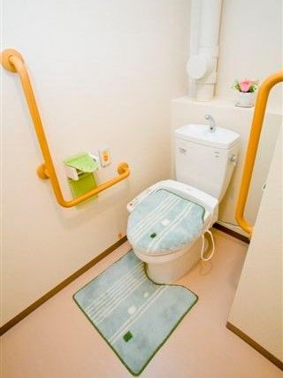 両サイドのオレンジの手すりと呼び出しコールが付いた居室トイレ。グリーンのトイレカバーが付けられ、トイレ奥の飾り棚にはピンクのバラが置かれている。