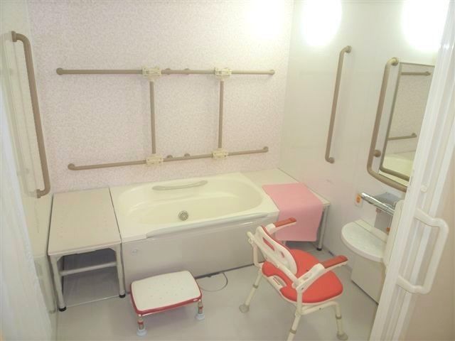 ３面に手すりが設置された白を基調とした浴室。浴槽の両サイドにはベンチがあり、赤いシャワーチェアが置かれている。