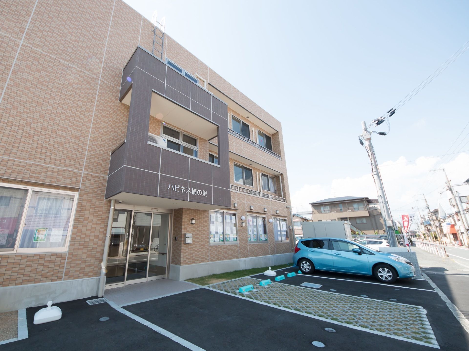 ブラウンを基調とした３階建ての建物になっている。施設前に車を停めるスペースがあり、車が１台停まっている。