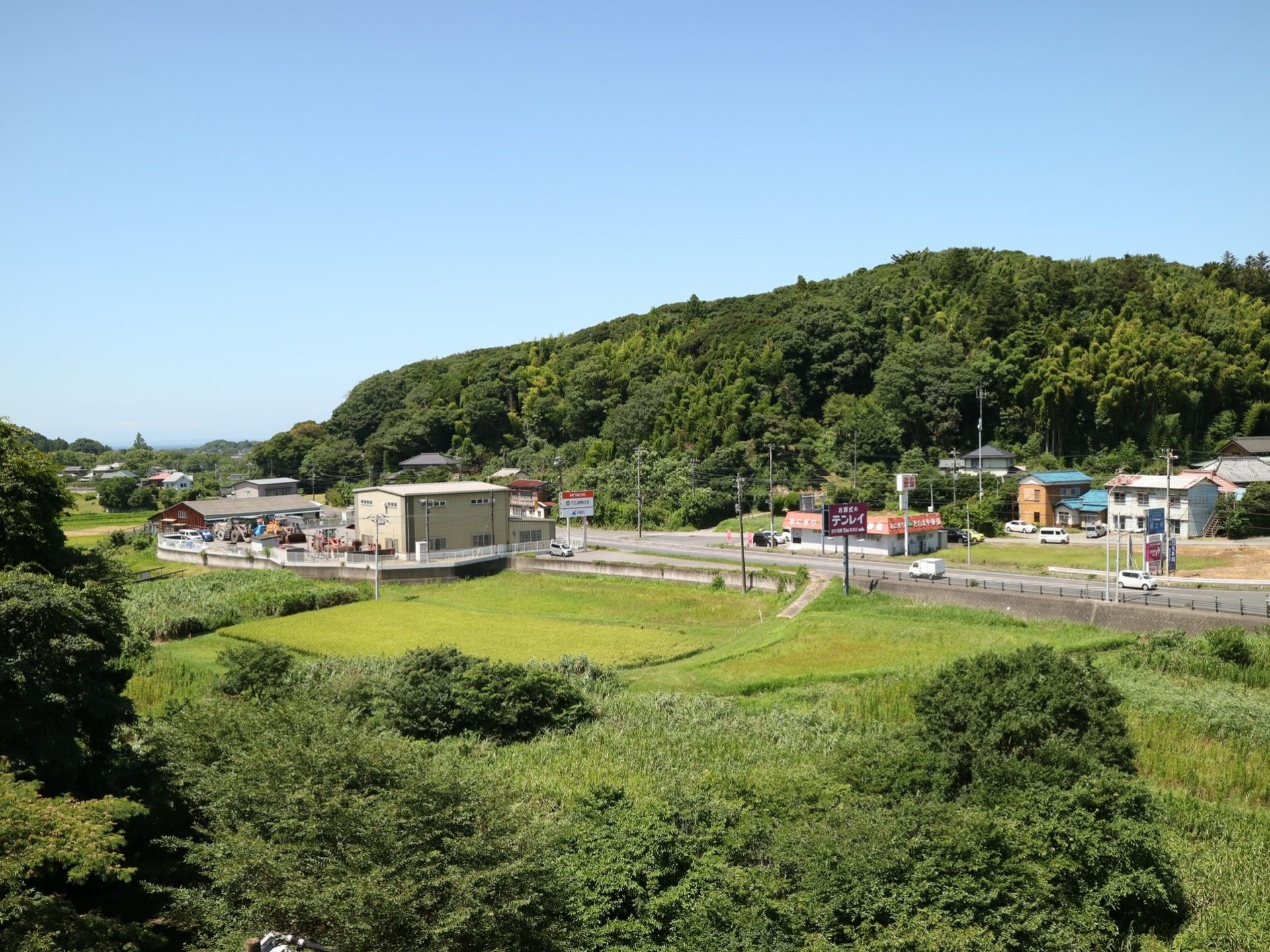 車が走っている大きな通りの周囲に民家や店舗がいくつか見える。その周囲には田畑や山など豊かな自然か広がっている。