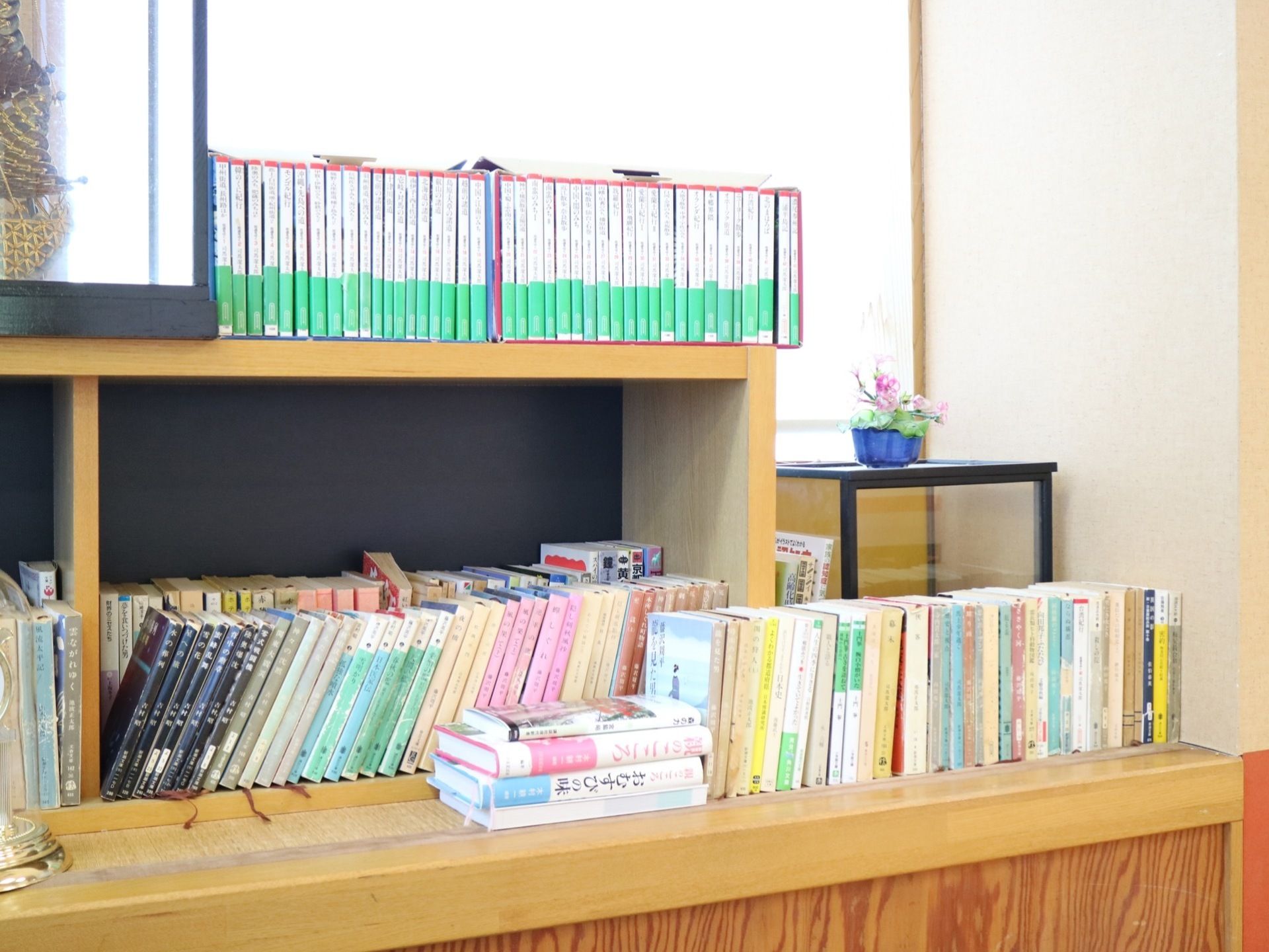 木製の棚に文庫本サイズの本がたくさん並べられている。棚の前や上にもさまざまな種類の本が積み重ねられている。