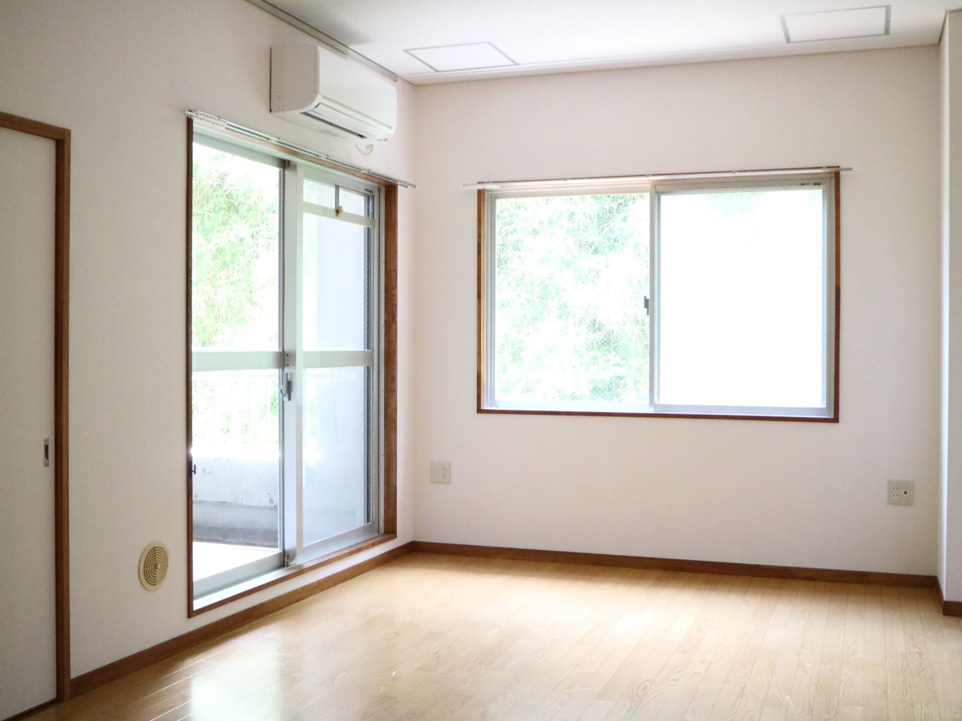 窓が２つ設置された明るい個室の部屋である。掃き出し窓の上にエアコンが１台取り付けられている。床はフローリングになっている。