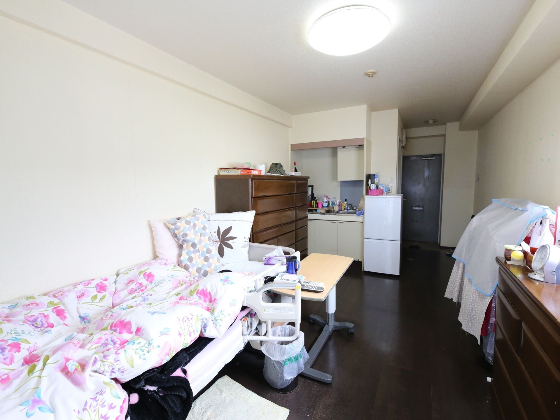 居室のベッドには転落防止柵がついている。冷蔵庫や衣類タンスが置かれている。パイプにかかっている衣類には布がかけられている。