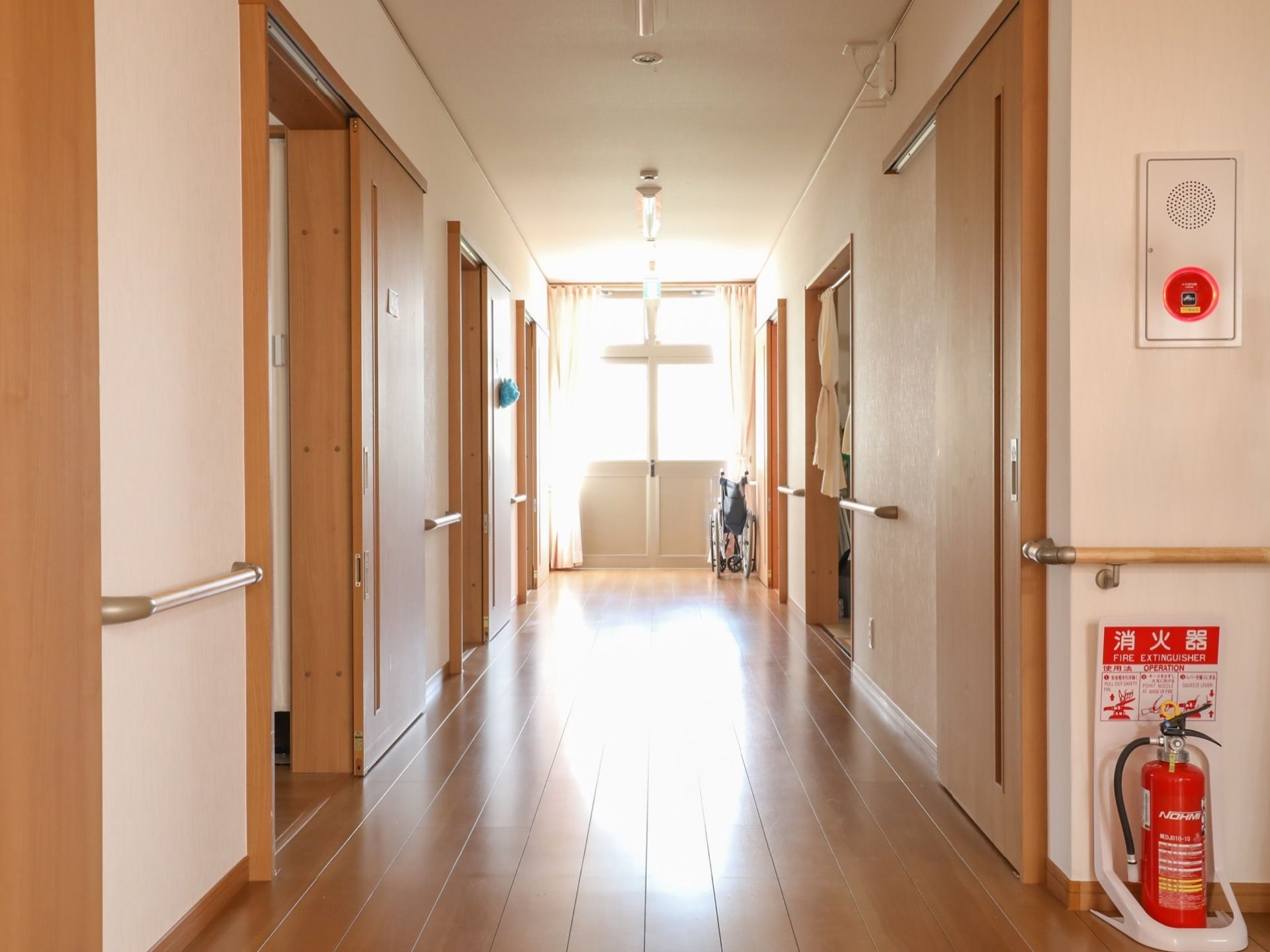 廊下には各居室のドアがあり、壁部分には手すりが設置されている。隅に非常用ベルや消火器が置かれている。