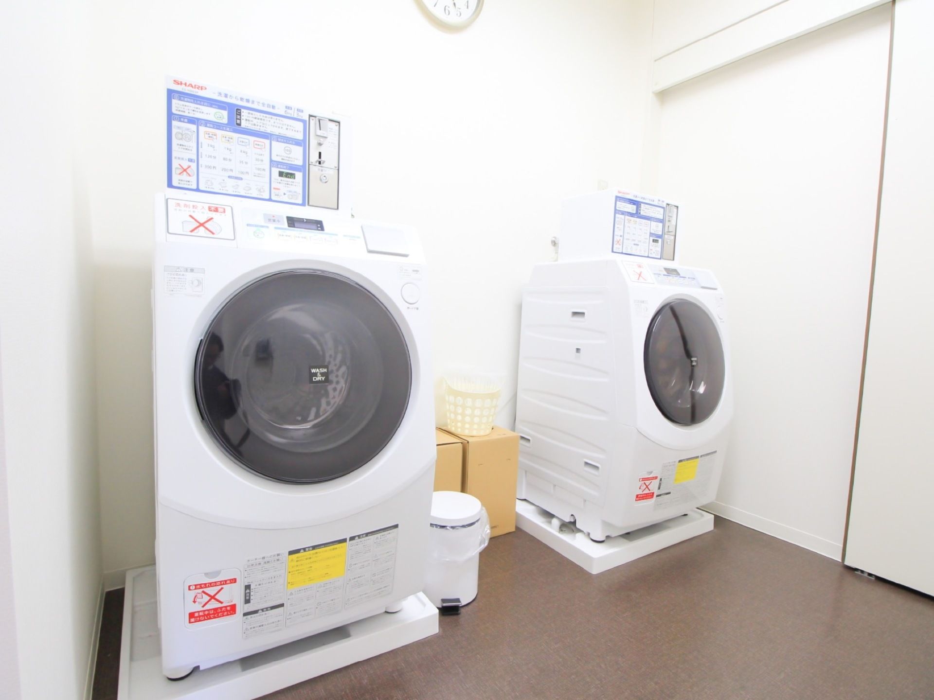 白を基調とした清潔感あふれるランドリー室で、ドラム式洗濯機が２台設置されている。壁には、時計が掛けられている。