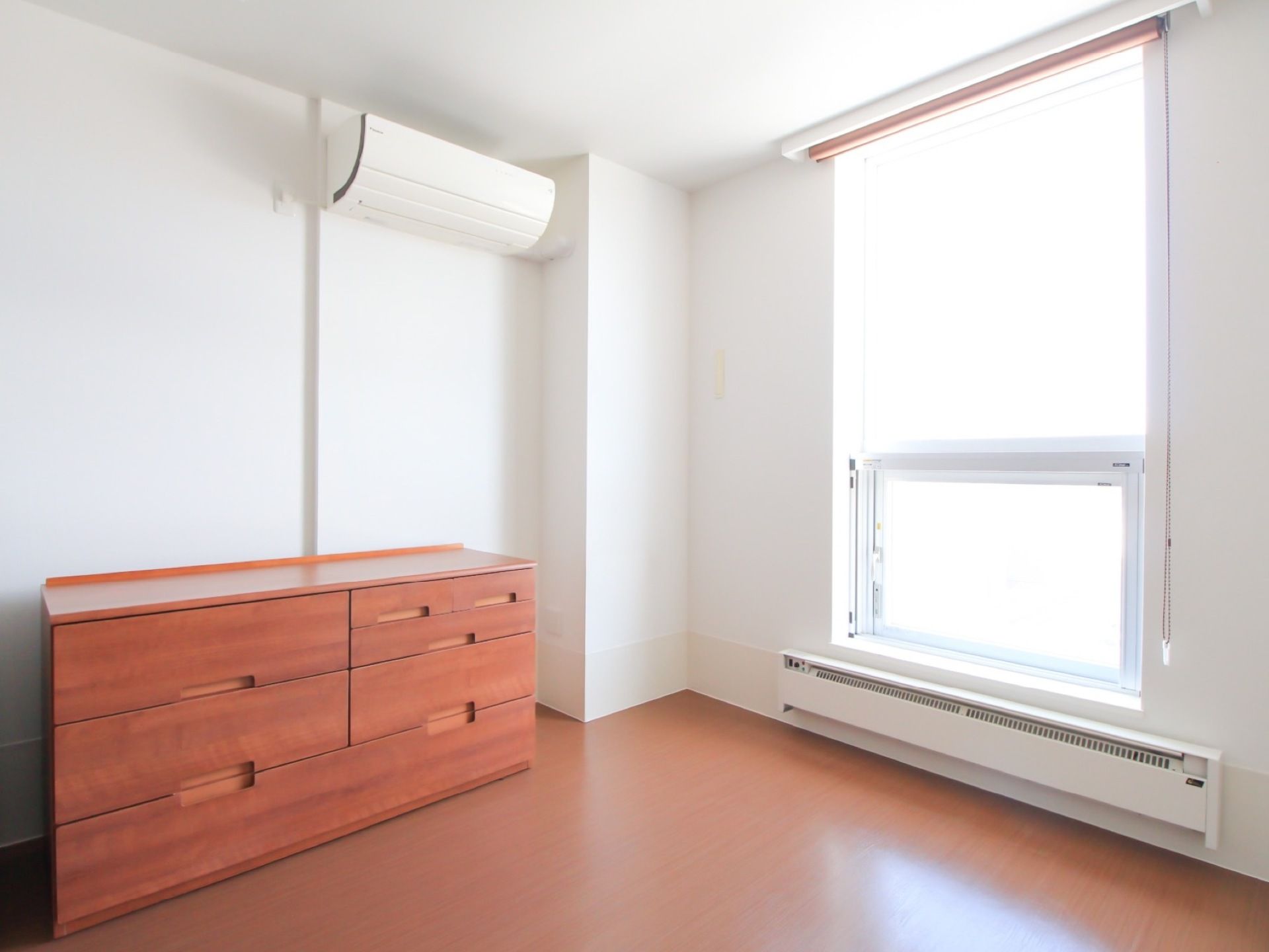 エアコンや暖房が完備されている部屋で、大きな窓にはロールカーテンが取り付けられている。壁際に収納棚が置かれている。
