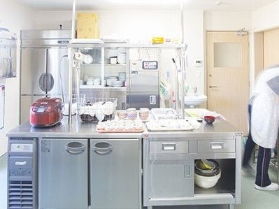 大き目のキッチンに炊飯器や業務用冷蔵庫が写っている。右側に調理している人がいる