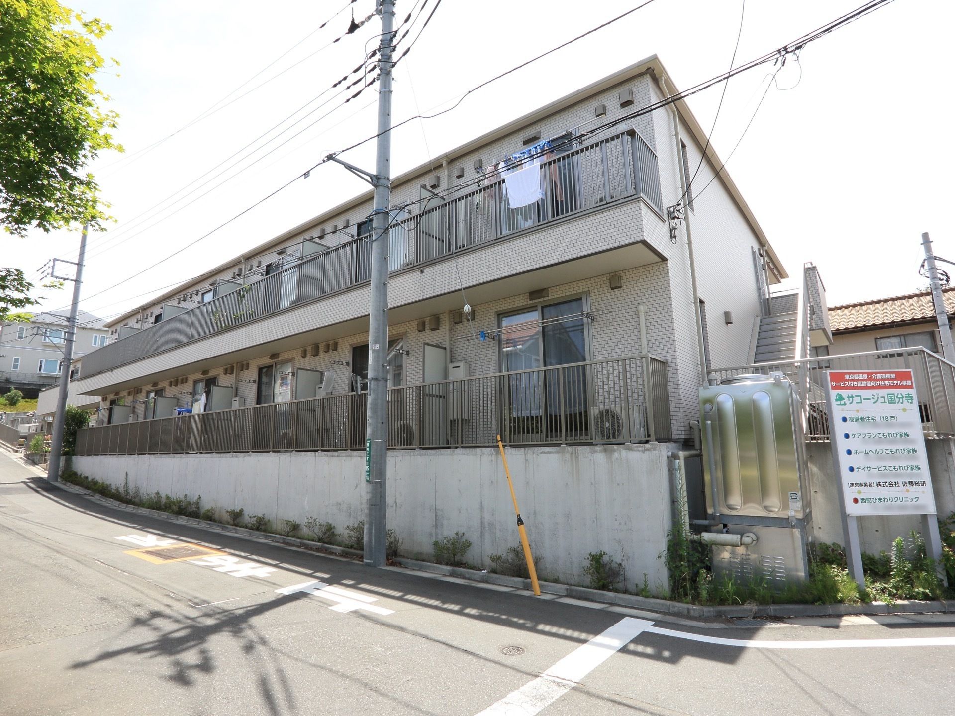 道路から高い敷地に建つ２階建ての施設である。各居室にはバルコニーがあり、洗濯物を干すスペースがある。避難階段が建物外部にある。
