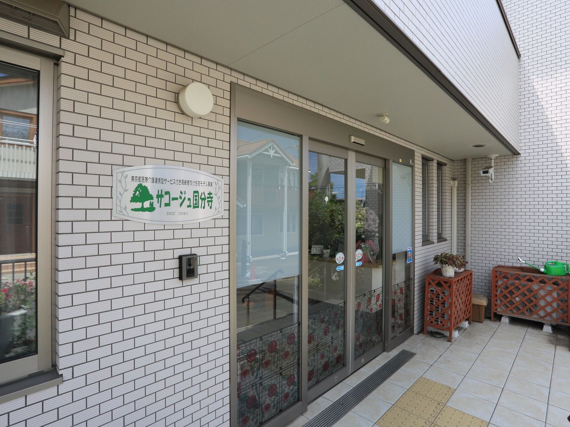 玄関は自動扉になっている。玄関周辺を映す防犯カメラが設置されている。施設名を記した看板が掲示されている。