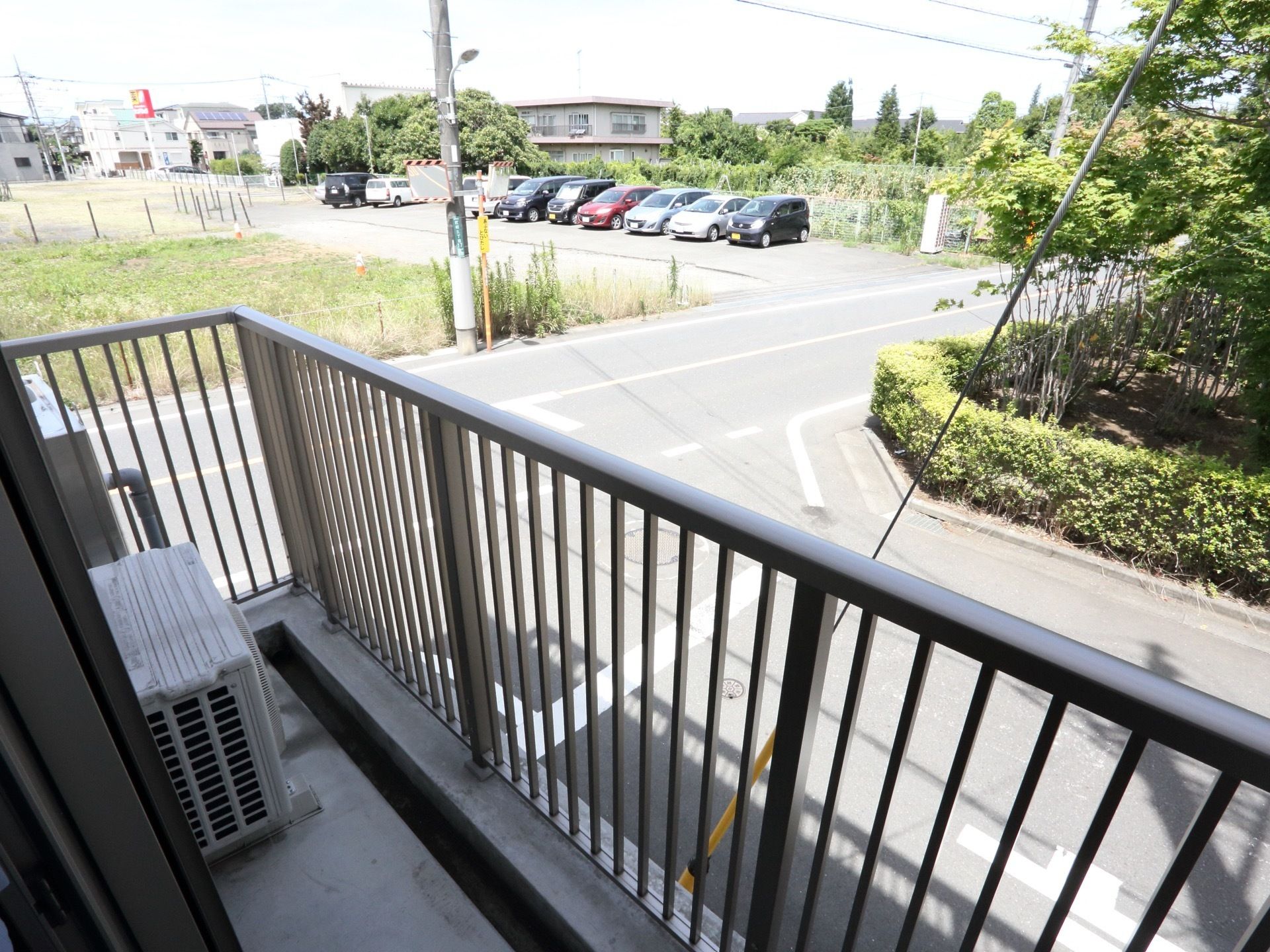 ２階の居室のバルコニーから見た近隣風景である。目の前には木々が茂っている。道路を挟んで駐車場がある。その隣は空き地となっている。
