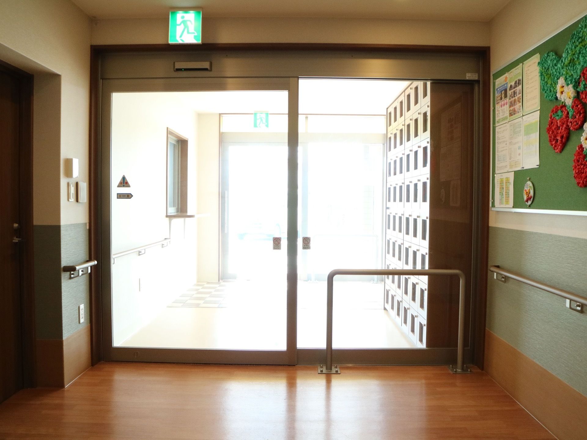 風除室のある施設の玄関部分。下駄箱の周辺に手すりが取りつけられている。そのほか、壁面に掲示板が設置されている。
