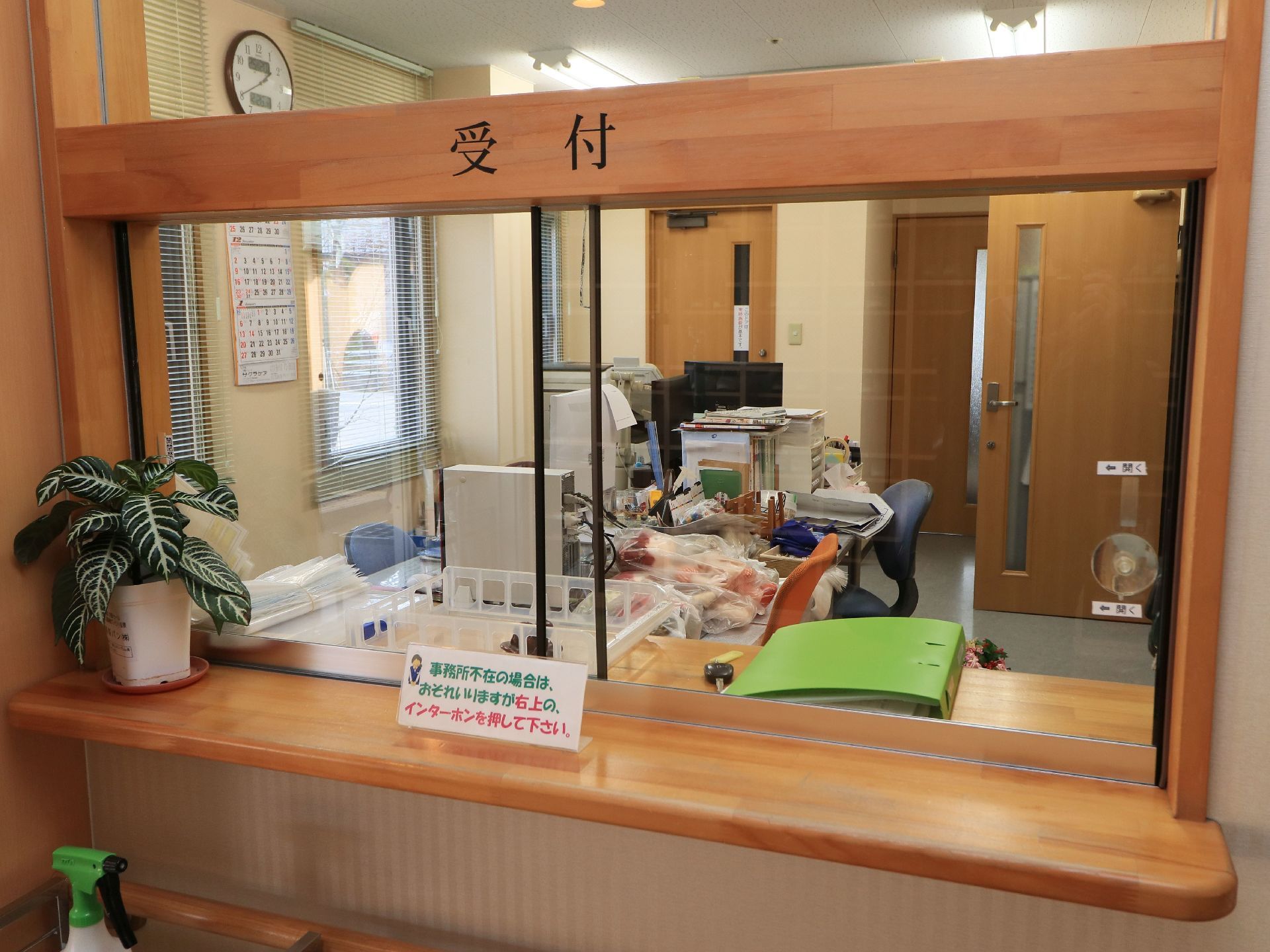 施設の風除室に受付が設けられおり、観葉植物がカウンターの上に置かれている。奥には、２つの事務机と椅子が配置されている。