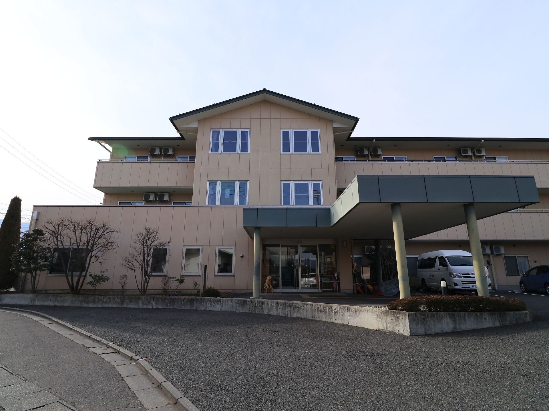 正面玄関前に車寄せが設けられている施設である。３階建ての建物には、窓が多く設置されている。建物前に駐車スペースが用意されている。