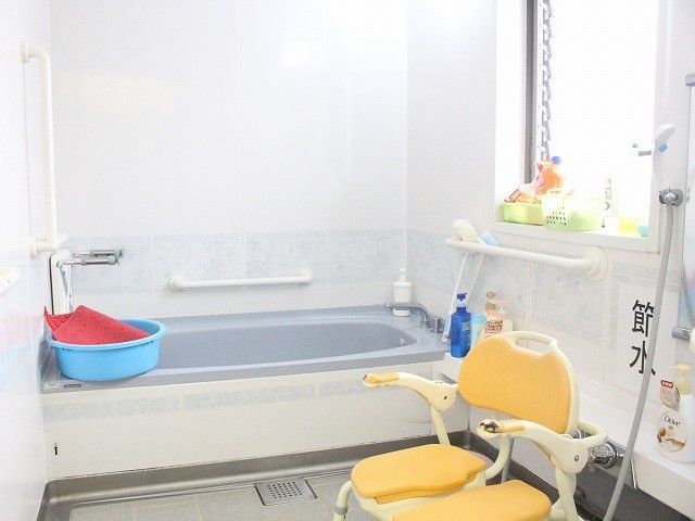 シャワーチェアや手すり、洗面器などが置かれた明るい浴室。「節水」の張り紙。