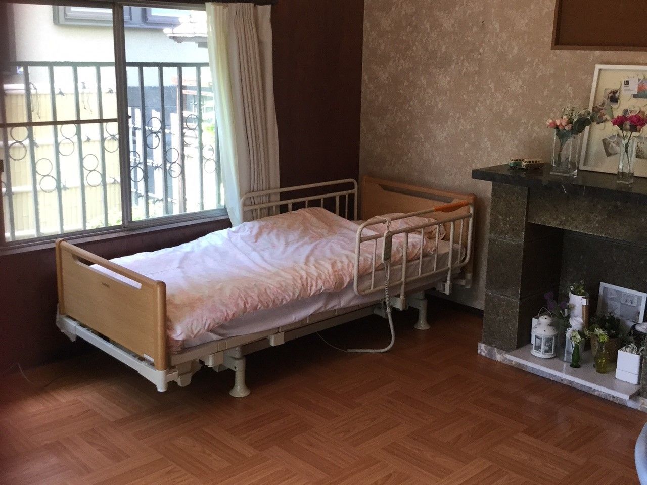 窓際にベッドが置かれたフローリングの部屋。右側には暖炉風のスペースがあり、花などが飾られている。