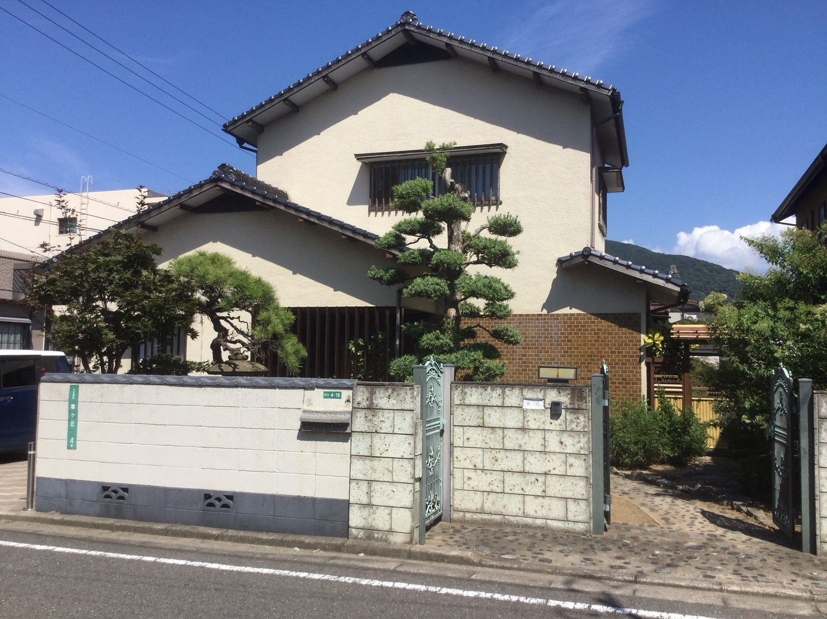 白いブロック塀で囲まれた木造２階建ての民家風の建物。庭先には多くの木も植えられている。