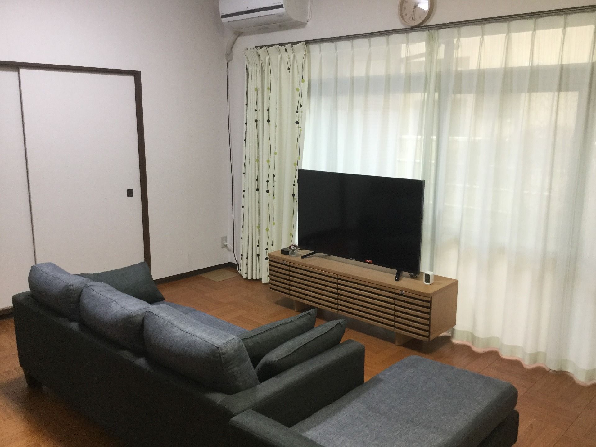 壁一面が窓となっていて明るいリビング。大型のテレビが置かれたテレビ台の向かいにはソファが置かれている。