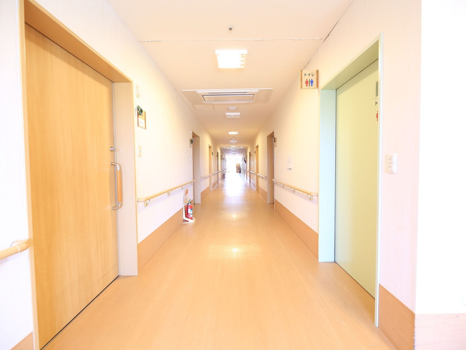 廊下突き当りの窓から陽光が差し込んで、床面に反射している。廊下脇にある居室の扉は、引き戸式である。廊下中央の壁際に消火器が置かれている。