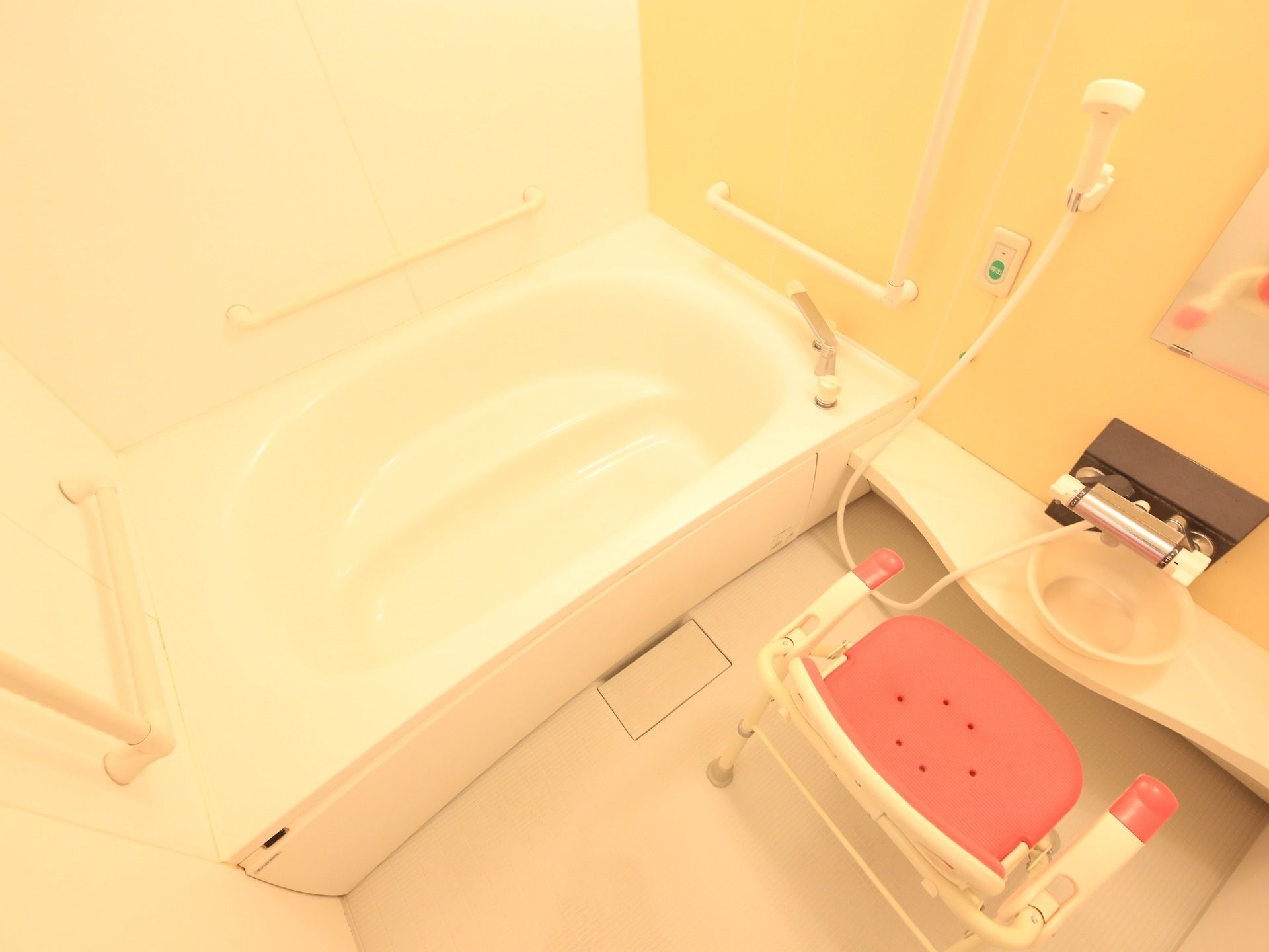 洗い場にシャワーとバスチェアー、洗面器が置かれている。白い浴槽の前方、後方、側面に手すりが取り付けてある。壁にナースコールがある。