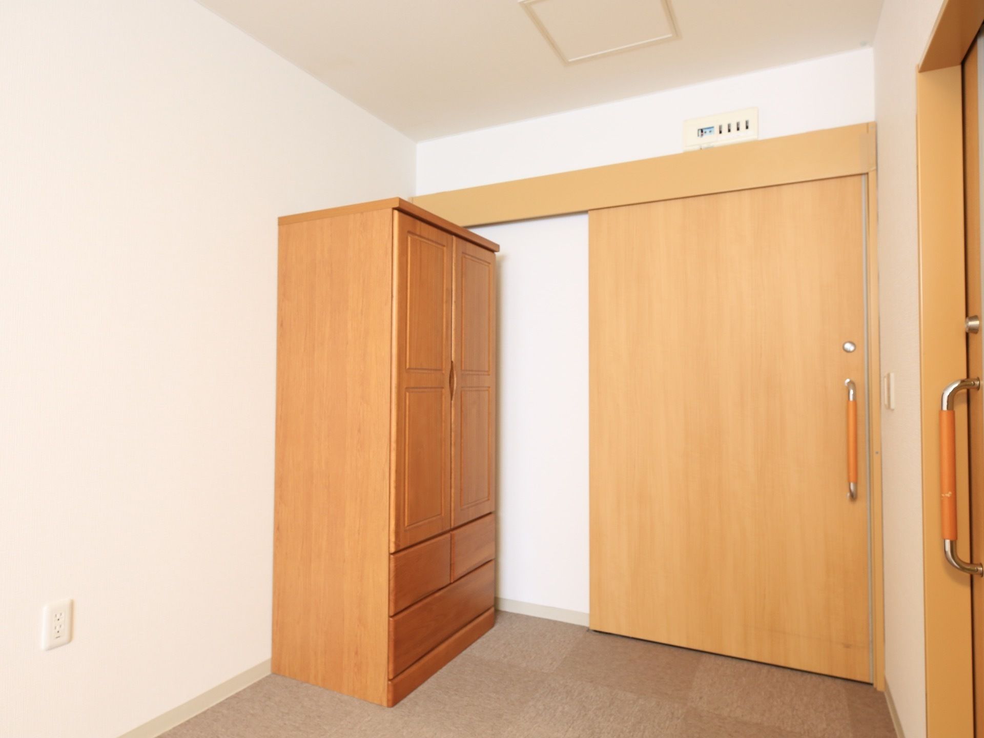 居室の入口脇に木製の収納タンスが置かれている。入り口のドアは、引き戸式で大きな取っ手が付いている。タンス横の壁に、電気コンセントがある。