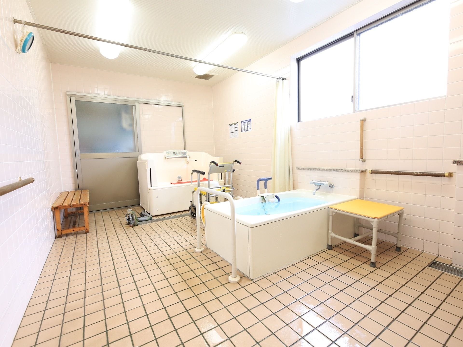 浴室がカーテンで２つに区分できる。奥に機械浴設備が置かれている。浴槽前に浴室用車椅子がある。手前に浴槽淵に浴用手すりの付いた一般浴槽がある。