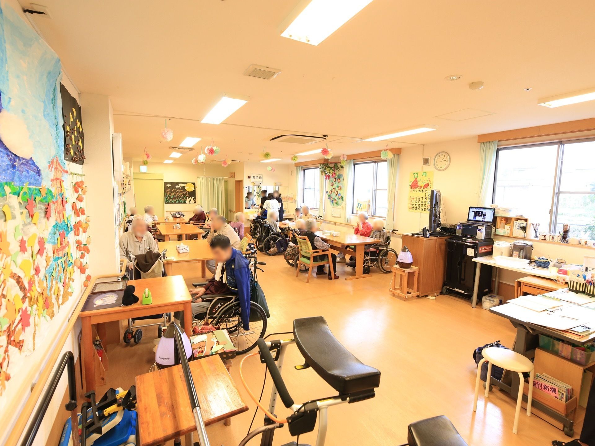 腰高窓が多くある部屋である。手前に機能訓練用器具が置かれている。奥に、テーブルと椅子がセットされ、数名の入居者様やスタッフが歓談中である。