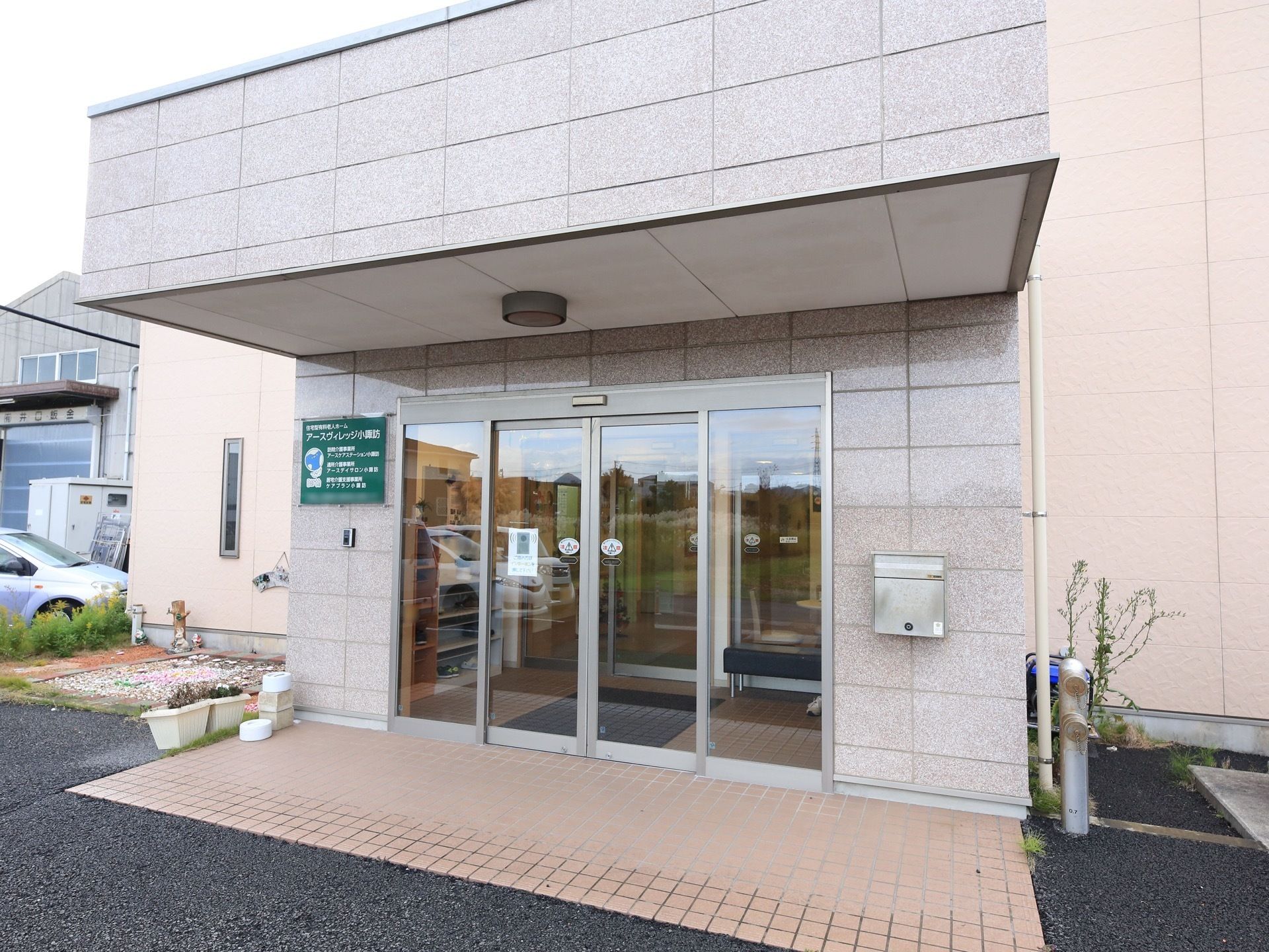 ガラスドアの玄関上には、ひさしが突き出している。ドアの左側の壁に緑地のプレートに施設名が記入されている。右側には、ポストが取り付けてある。