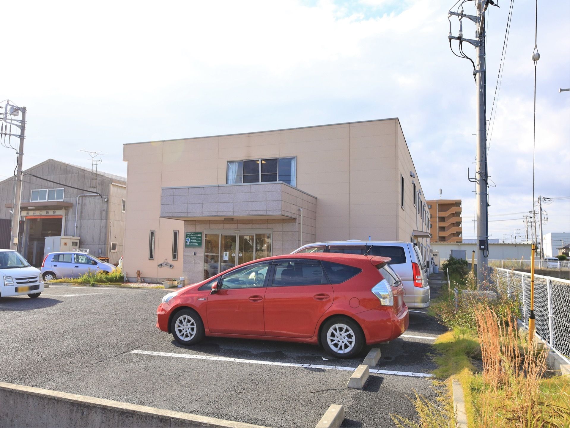 ２階建ての施設の前に駐車場が設置されている。アスファルト舗装がされ、白線で区分されている。施設敷地の境には、白いフェンスが張られている。