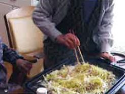 ホットプレートを使用して焼きそば作り。エプロンをして菜箸を握り調理に当たる男性の手元。