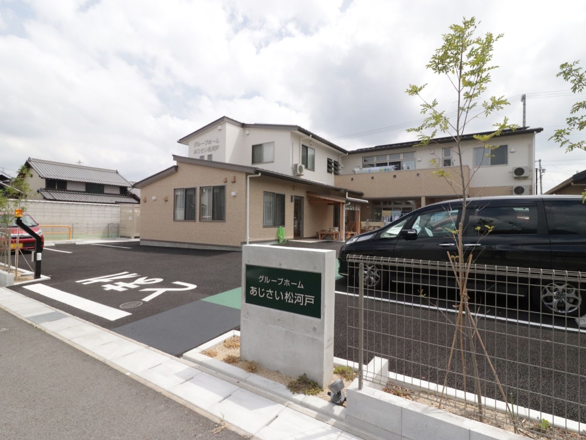 道路に面して、駐車場が広がり、その奧に２階建てのＬ字型の施設が建っている。駐車場の周囲にフェンスが廻らされ、植栽が施されている。
