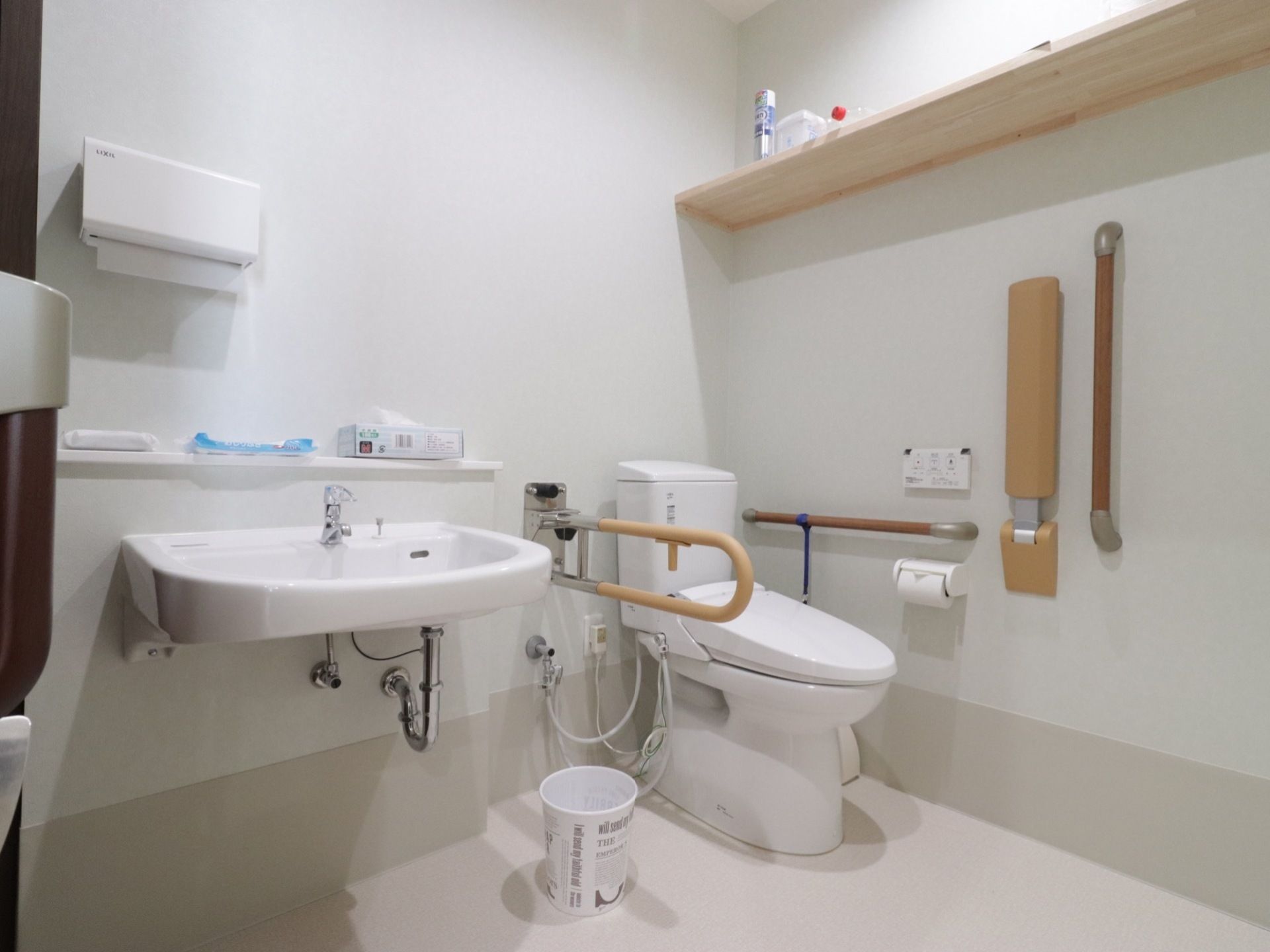 広いトイレにタンク付きの洋式便器が据えられており、さまざまな形の手すりが設置されている。掃除用具や衛生用品が置かれている。
