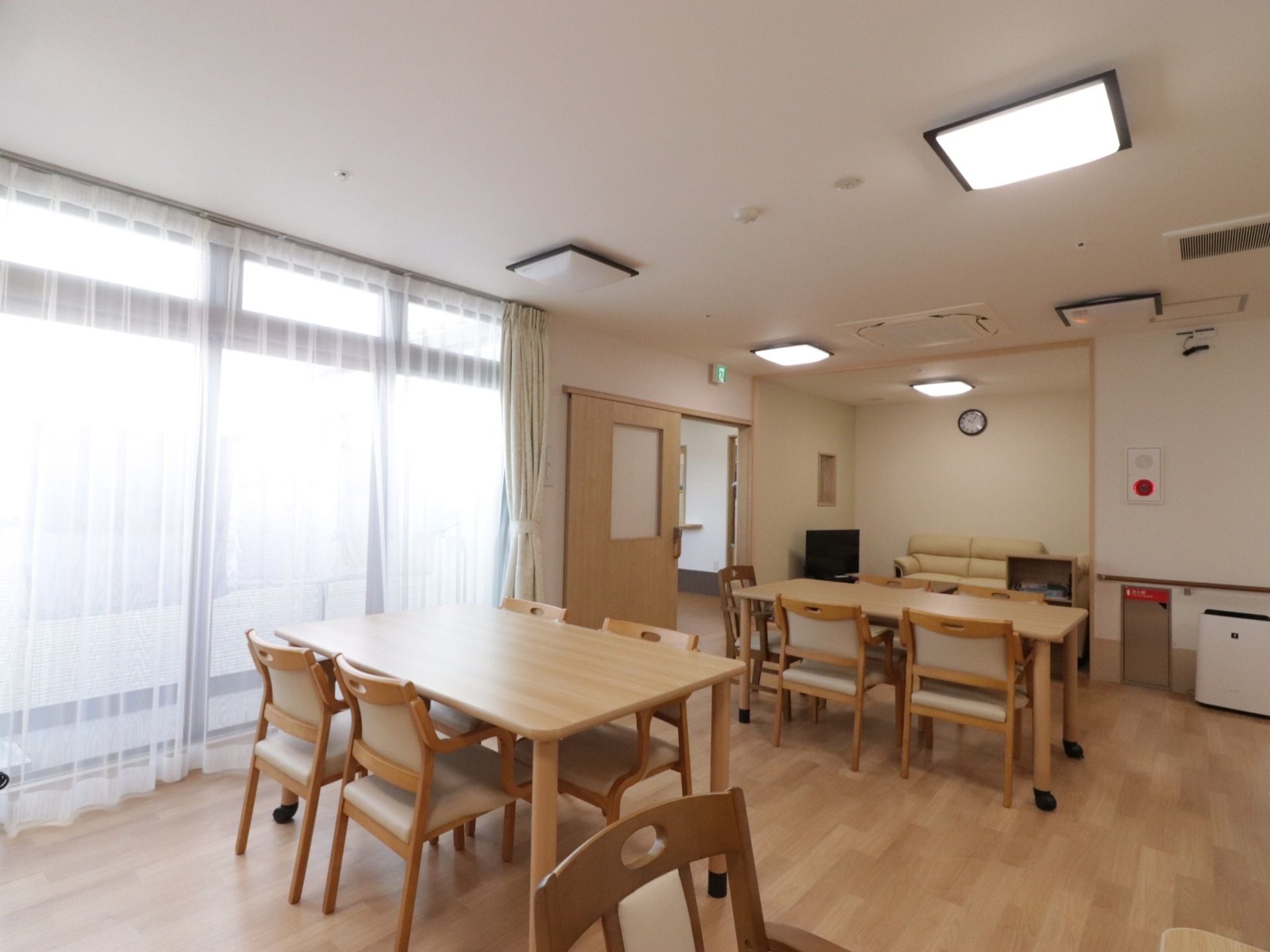複数のダイニングセットが間隔を空けて配置されている食堂。奧に、ソファやテレビが置かれた畳スペースが見える。