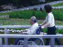 車いすに乗った高齢の男性がスタッフの介助を受けながら、外出を楽しむ風景。