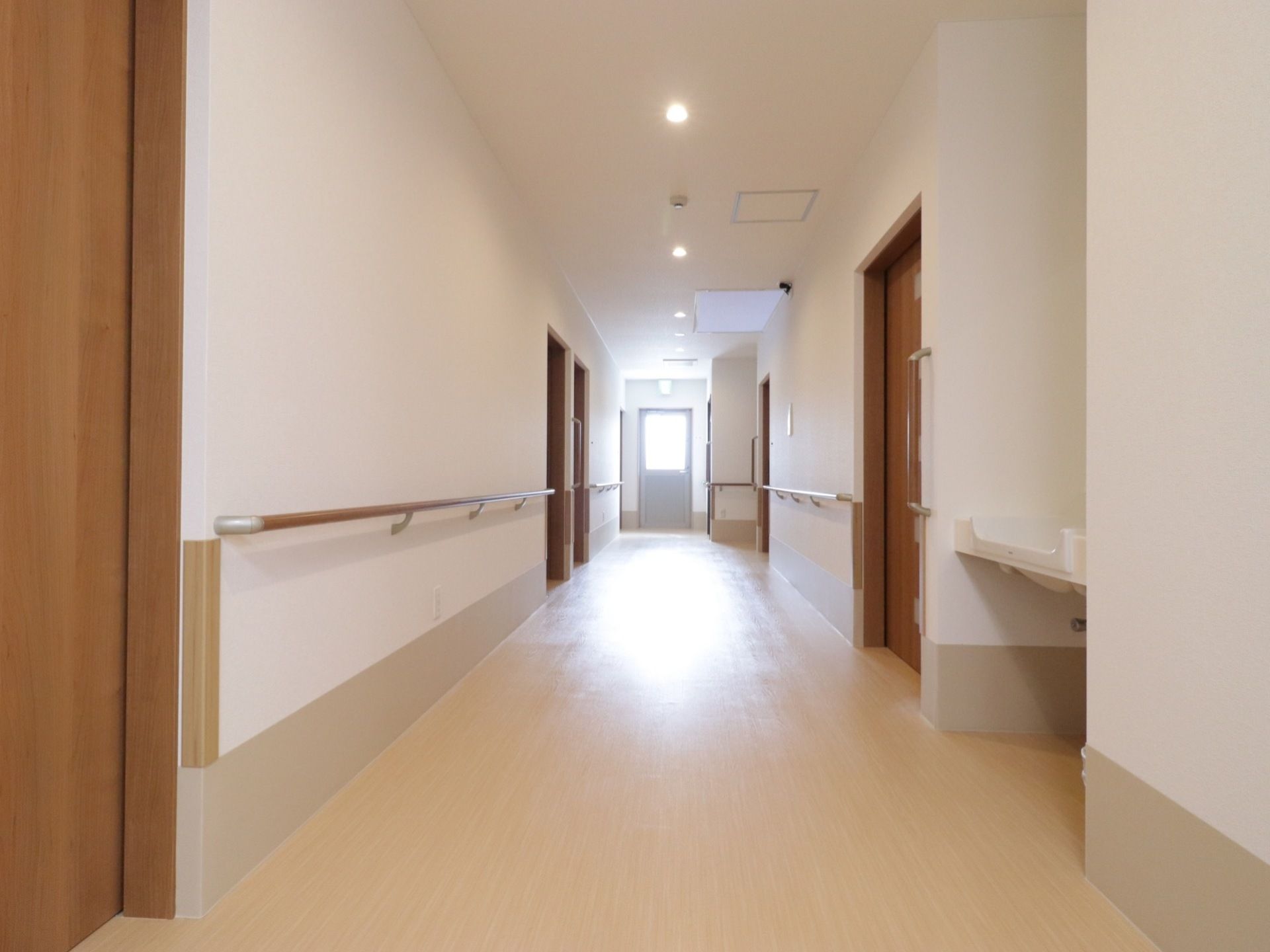 フローリングの廊下が伸びており、両側に居室が並んでいる。壁には手すりが設置されている。突き当たりに窓があり、明るい。