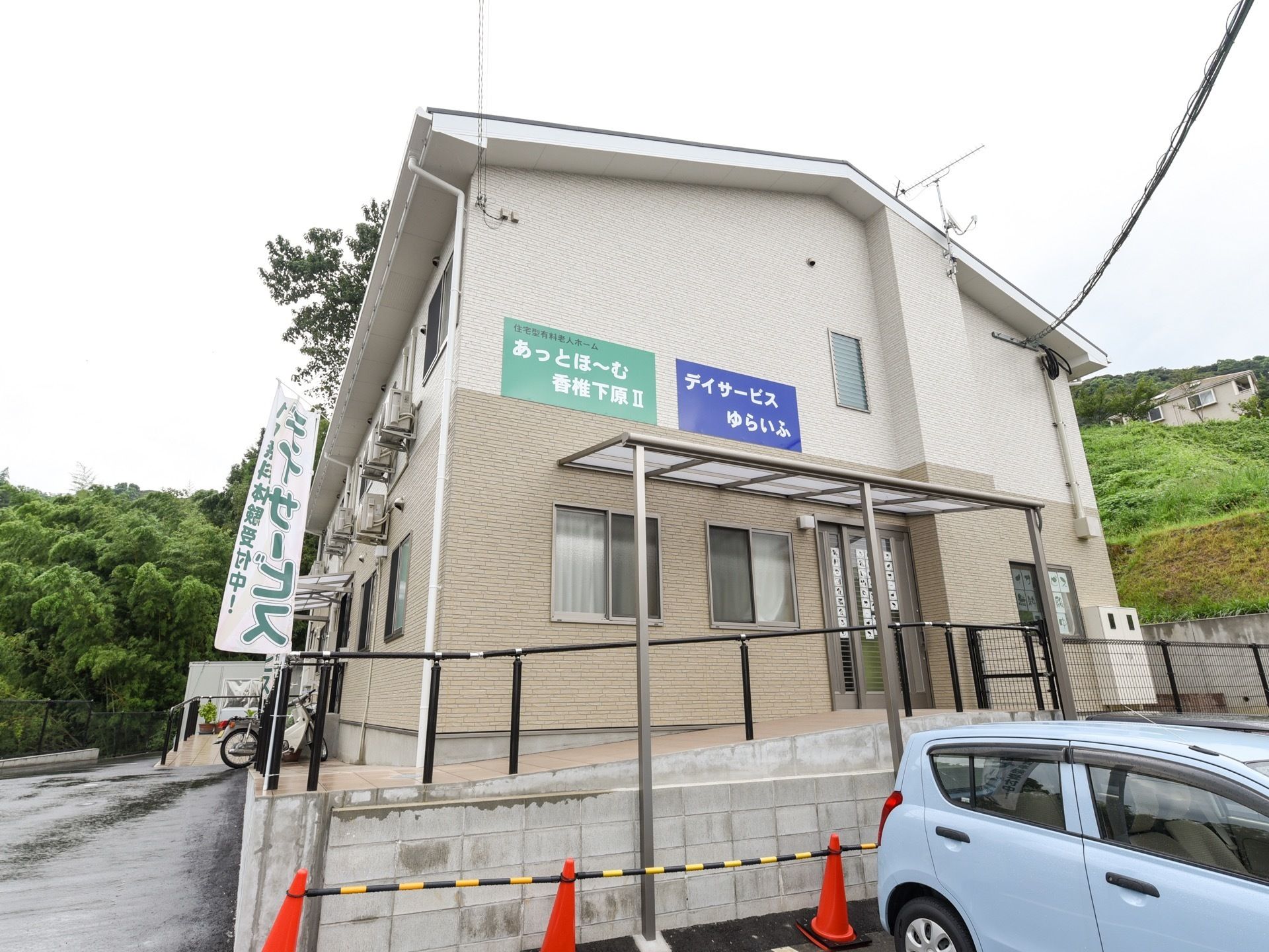 外壁がベージュ色の２階建ての建物である。デイサービスが併設されており、青色の看板に白色の文字で名称が表示されている。