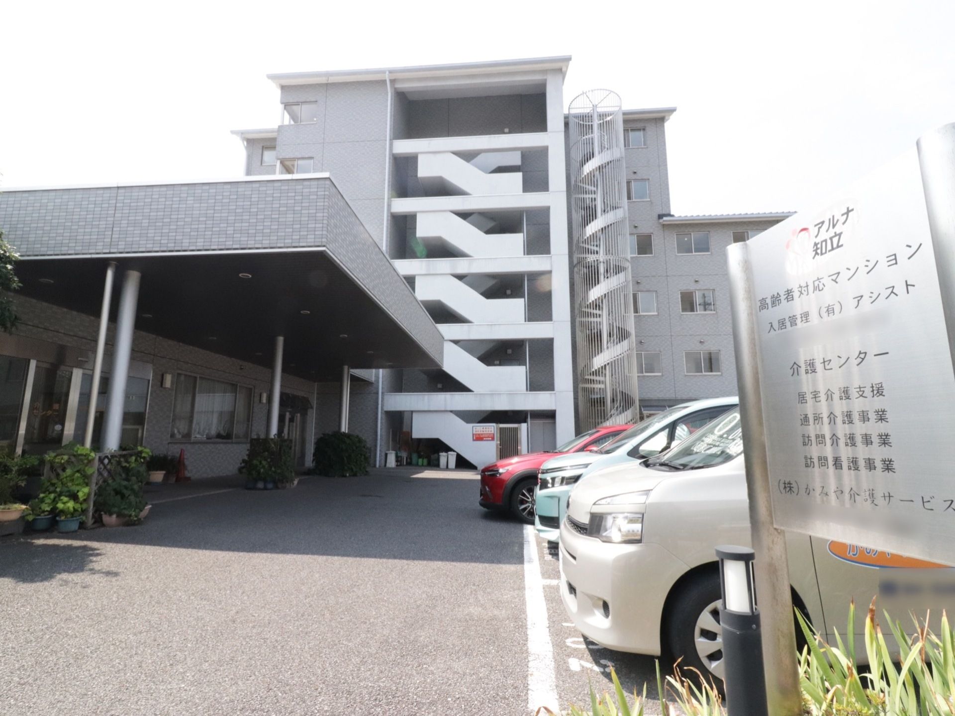敷地の入り口に施設名などが書かれた銀色の看板が立っており、その奥が駐車場になっている。建物入り口には大きなひさしがついている。