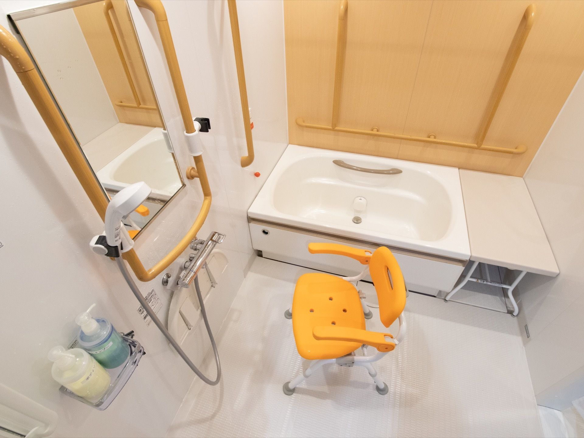 一面の壁に明るいブラウンを採用した浴室である。浴槽のすぐ隣に台を備え、横側の壁には縦と横に手すりを設置している。