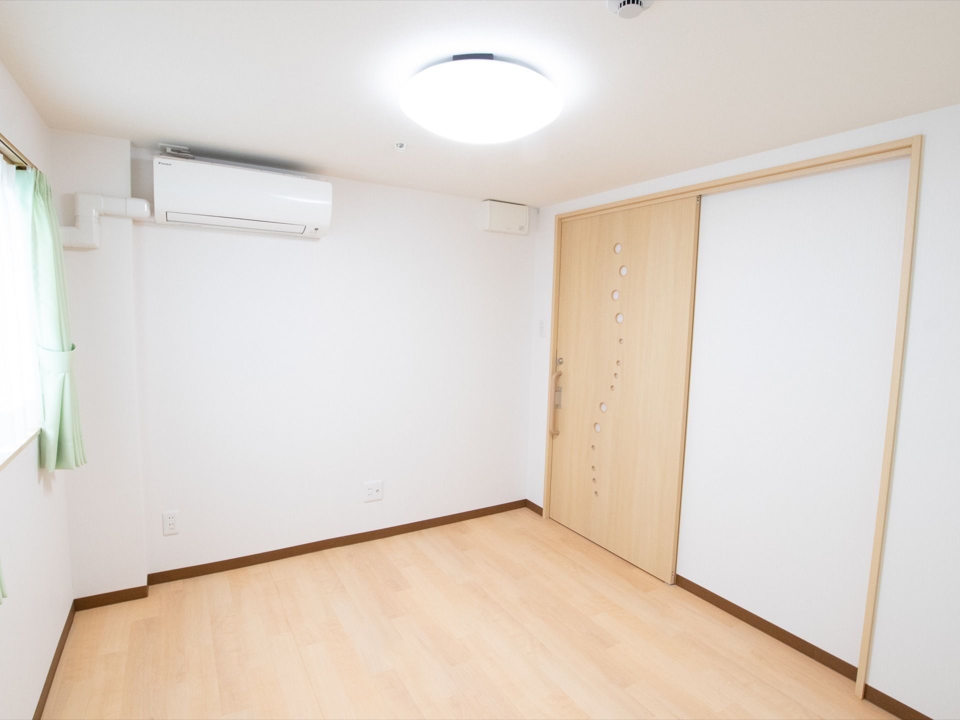 白木調のドアとフローリングの居室である。天井に円盤型の照明を設置し、空調設備を整えている。窓にはレースと水色のカーテンを掛けている。