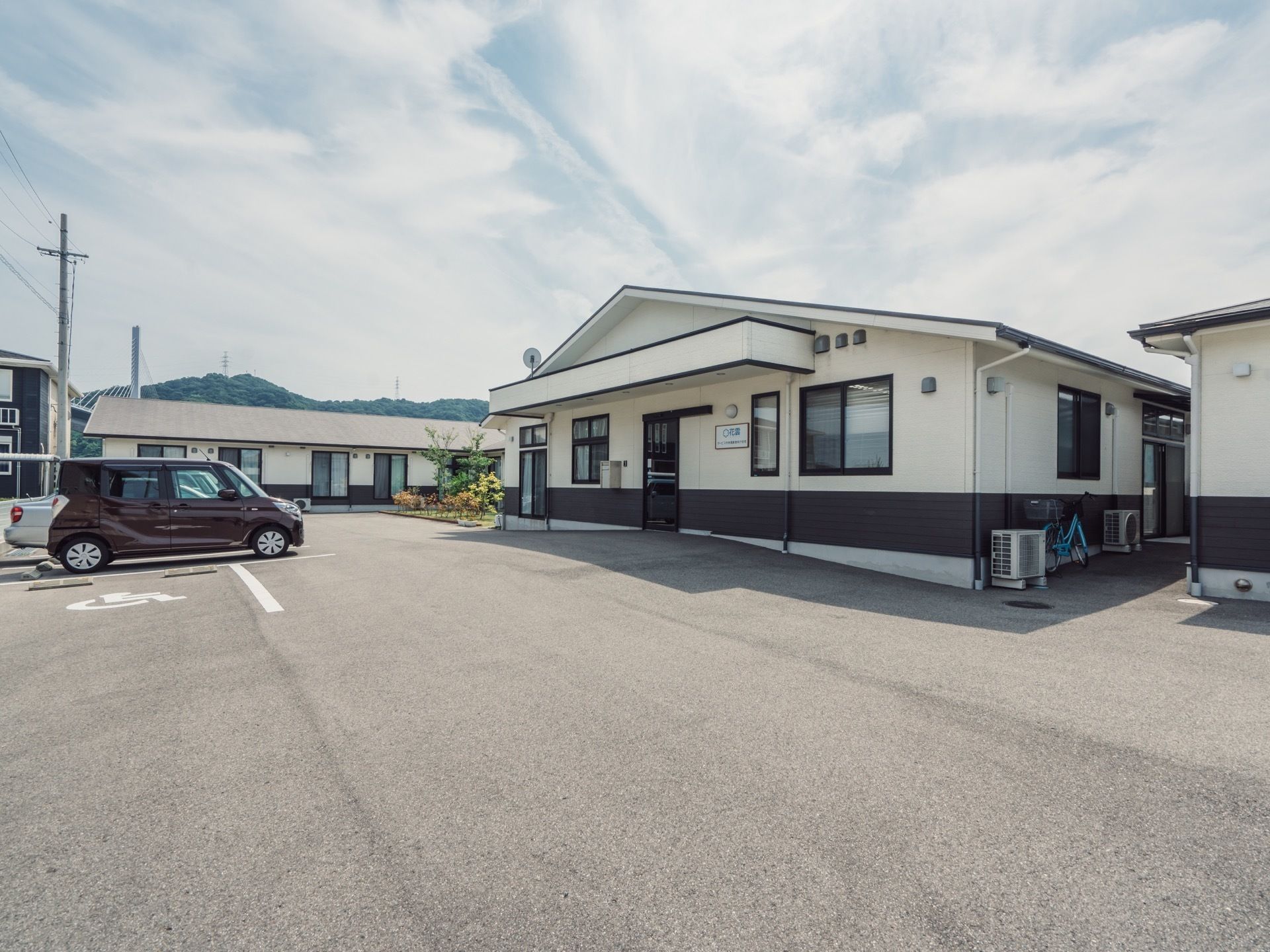 広々とした駐車場が施設の左側にあり、数台の車が並んでいる。建物の後方に、緑の山並みとスカイブルーの空が見える。
