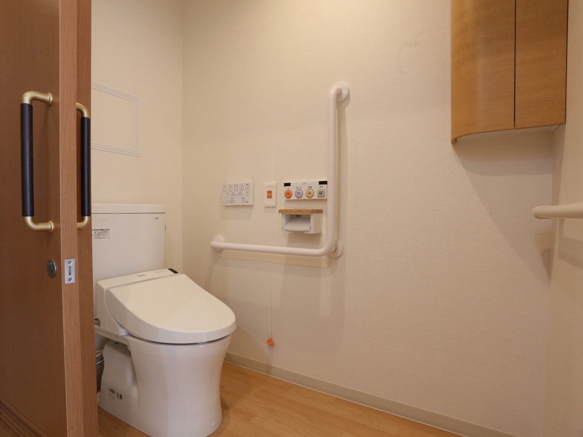 白い壁の個室トイレで、入り口はスライド式の扉が使用されている。壁にL字型の手すりが取りつけられている。
