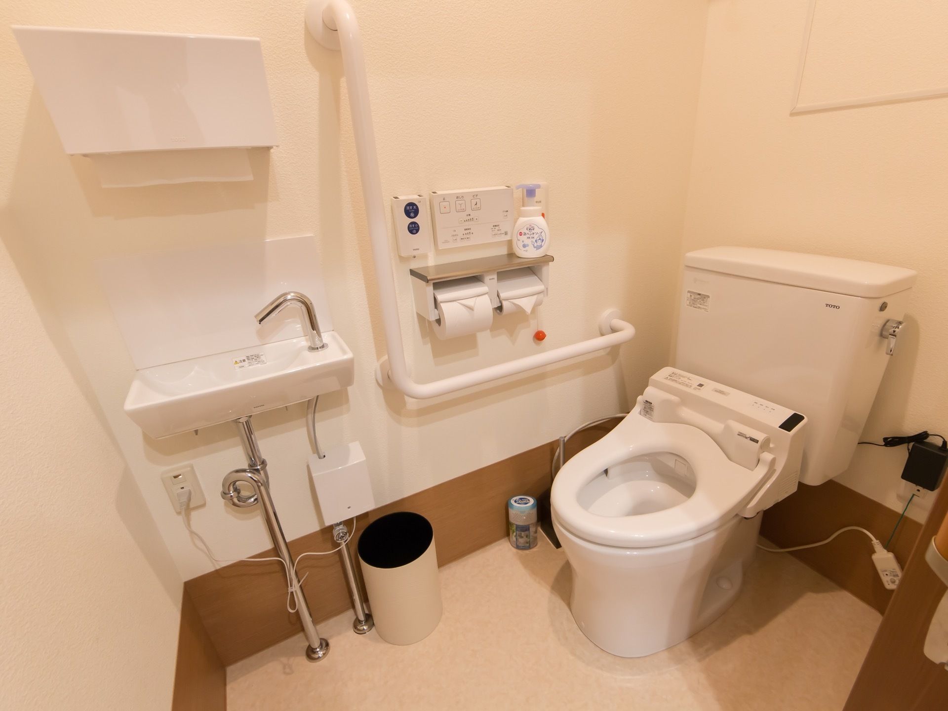 温水洗浄便座の備えられた便器と洗面台のある個室のトイレがある。壁には手すりが設置され、ペーパーホルダーが付いている。