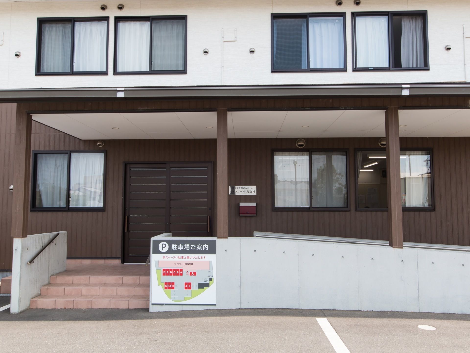 建物の周囲にアスファルトで舗装された駐車場が広がっている。駐車場から玄関へ続く緩やかな傾斜のスロープがある。