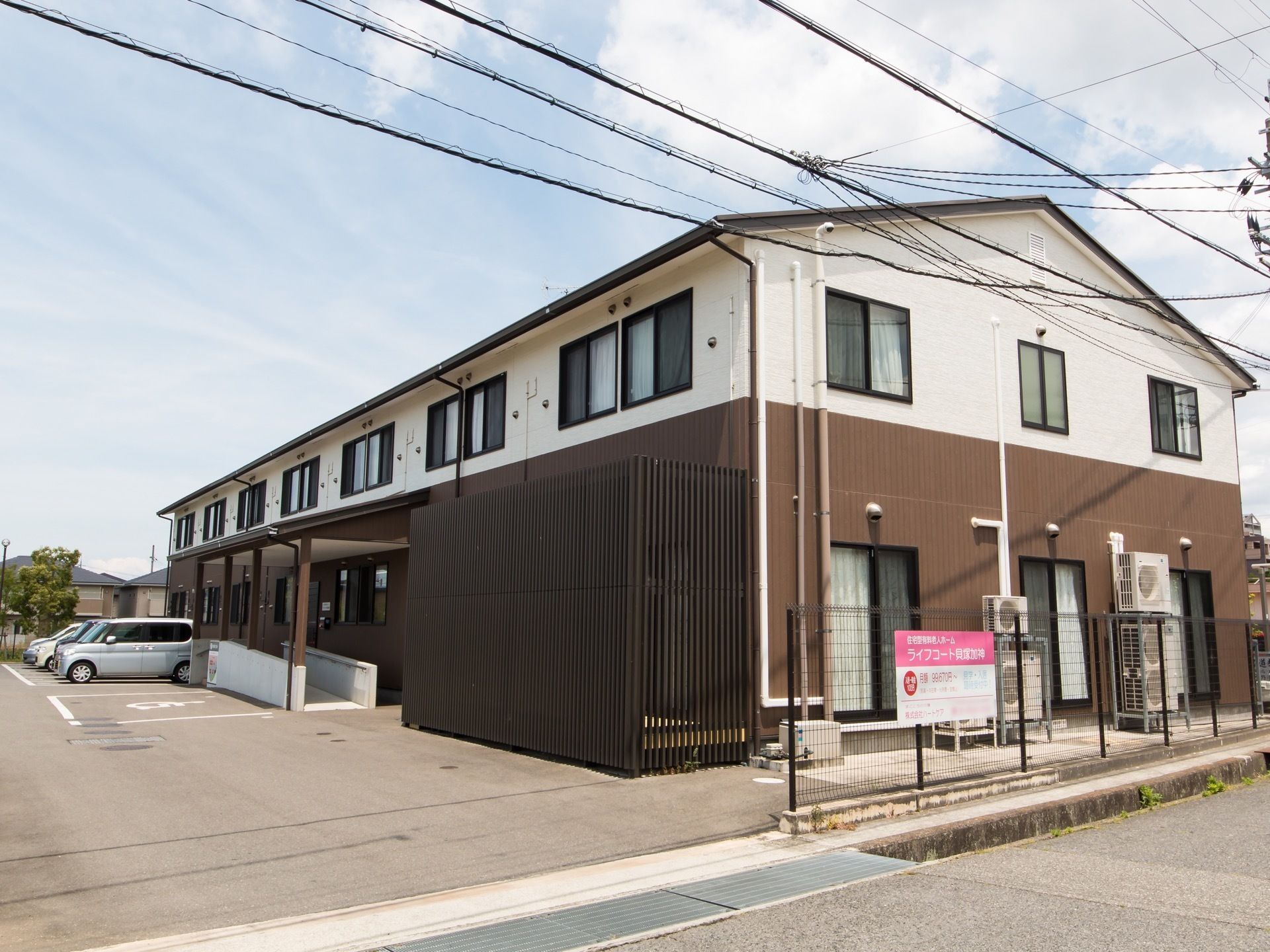 横幅の広い2階建ての施設が建っており、前面にはアスファルトで舗装された駐車場が広がっている。フェンスの手前には道路が見える。